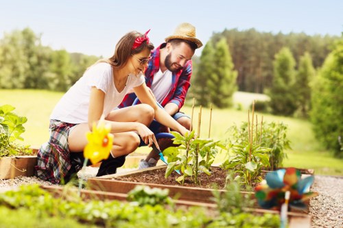 A woman and a man are working together in a well-maintained backyard garden, planting and tending to young vegetable plants in a raised bed with rich soil. The garden features a lush, green lawn in the foreground and is bordered by other garden beds and decorative elements like a small yellow garden ornament and a colorful bird bath. In the background, there are tall, leafy trees under a bright blue sky with some clouds, indicating a sunny day. The scene captures outdoor gardening activity, with the gardeners appearing engaged and attentive to their work, highlighting sustainable gardening practices. The environment suggests a suburban setting, possibly in Ilford, with an emphasis on organic growth and eco-friendly landscaping, aligning with the ethos of recyclability and sustainability promoted by Landscapers Ilford. The natural light enhances the vibrant greenery and the overall peaceful atmosphere of the garden, reflecting a well-organized outdoor space dedicated to eco-conscious gardening efforts.