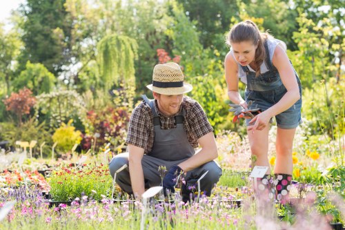A garden scene in a residential outdoor space featuring a well-maintained lawn with dense, green grass in the foreground, bordered by flower beds filled with vibrant, colorful blooming flowers and small shrubs. The garden has a backdrop of taller trees with leafy canopies providing shade and privacy. Two women are engaged in gardening activities; one is crouched down, wearing a straw hat, checked shirt, and gardening apron, tending to the flowers with gardening tools, while the other, dressed in a sleeveless top and denim shorts, is standing and holding pruning shears, appearing to prune or inspect plants. The garden has a mix of soil, paving stones, and grassy areas, creating a natural outdoor environment suitable for gardening maintenance and landscaping services. The lighting suggests a bright, sunny day, emphasizing the lush greenery and vibrant flower colours, reflecting a well-kept and cared-for garden space near Ilford, supporting local gardening and landscaping expertise by Landscapers Ilford on their website.