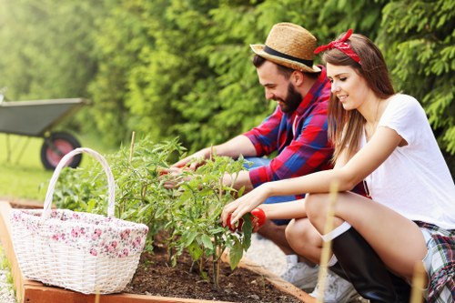 A man and a woman gardening together outdoors in a residential garden, surrounded by lush green trees and shrubbery. They are crouched down beside a raised flower bed made of wood, tending to young plants with green foliage. The man, wearing a straw hat and a red and blue plaid shirt, is reaching into the soil, while the woman, with long dark hair tied with a red headband and dressed in a white t-shirt and plaid skirt, is carefully handling a plant. A white wicker basket with pink floral patterns is placed on the edge of the flower bed, filled with gardening tools or flowers. In the background, a lawn area and garden furniture are visible, with natural sunlight illuminating the scene. The setting suggests a well-maintained garden dedicated to planting and outdoor gardening activities, aligned with professional gardening services such as those offered by Landscapers Ilford in the local area, including Ilford and the IG postcode district.