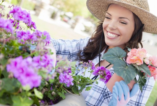 A smiling woman in a wide-brimmed straw hat and light blue gardening gloves tends to an array of vibrant flowering plants in a well-maintained outdoor garden space. The garden features a lush green lawn with dense hedge borders in the background, alongside a variety of flowering plants including purple, pink, and orange blooms, which add natural colour variation. The woman appears to be carefully inspecting or pruning the plants, with her hand near the flowers, suggesting active garden maintenance. The scene is brightly lit, indicating a sunny day with natural daylight illuminating the garden layout, which includes flower beds filled with soil and areas of dense grass. The garden’s structure shows a combination of lawn, flower borders, and possibly a paved area or decking in the background, creating a neat and inviting outdoor environment. This setting aligns with professional gardening or landscaping services aimed at maintaining vibrant and healthy outdoor spaces, such as those provided by Landscapers Ilford, serving properties in the Ilford area, within the IG postcode district across Essex and East London.
