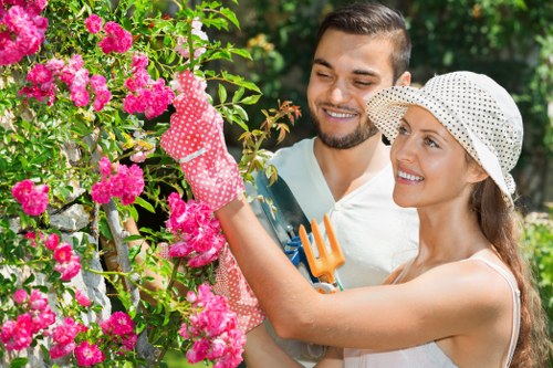 A photograph of a man and woman in a garden, with the woman wearing a white sun hat and pink gardening gloves, gently pruning a vibrant pink flowering shrub. The garden features a well-maintained lawn in the foreground, with lush green foliage and trees in the background. The scene is captured during daylight with natural sunlight illuminating the garden, showcasing a neat arrangement of plants and flowers, indicative of ongoing gardening or maintenance work by a professional landscaper in Ilford. The presence of gardening tools such as a small garden fork held by the woman highlights active outdoor landscaping and gardening efforts, supporting outdoor maintenance services offered by companies like Landscapers Ilford within the local area.