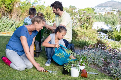 A family of four gardening outdoors in a well-maintained backyard with lush green grass, flowering plants, and shrubs. The mother, wearing a blue top, is kneeling on the grass, using a small gardening fork to tend to a young plant in the foreground. Next to her, a girl with a blue backpack is watering plants from a white container, with garden tools and a clay pot nearby. The father, dressed in a light shirt, and a boy in a striped shirt are standing behind, engaged in gardening activities amidst the vibrant garden beds that include various perennials and ornamental shrubs. The garden features a mix of natural soil and neatly edged flower beds, with a backdrop of a scenic view including hills and a partly cloudy sky, indicating a pleasant outdoor environment. The scene suggests regular outdoor maintenance and gardening, aligning with professional landscaping and lawn care services provided in the Ilford area, especially around postcodes IG1 and IG2, enhancing the garden's overall health and appeal.