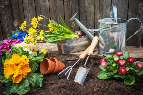 A garden scene featuring a freshly planted flower bed with rich, dark soil in the foreground, bordered by a weathered wooden fence in the background. To the left, there is a cluster of colorful flowers, including yellow primroses and pink primulas, with lush green foliage. On the right, potted plants with vibrant pink blooms are arranged nearby. Gardening tools such as a small hand rake, trowel, and watering can are laid out on the soil, indicating ongoing planting activity. A metal trough filled with yellow and white daffodils rests behind the tools, along with empty terracotta pots. The overall setting suggests a neatly maintained outdoor space suitable for gardening and landscape enhancement, consistent with services offered by Landscapers Ilford in the local Essex area. The scene is illuminated by natural daylight, highlighting the fresh and thriving garden environment, which is ideal for enhancing outdoor landscapes and lawn care in the Ilford region.