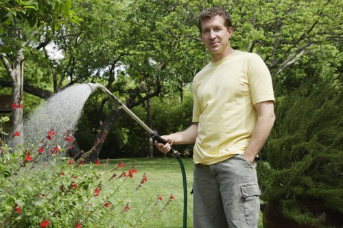 A man standing in a well-maintained garden during daytime, holding a garden hose with water flowing onto a bed of vibrant red flowers and green foliage. The garden features a neatly trimmed lawn, bordered by various shrubs and small trees, with a mix of grass, soil, and flower beds visible. In the background, tall trees with lush green leaves create a natural canopy, and a dense hedge runs along the perimeter, providing privacy. The sun illuminates the scene with soft, natural light, highlighting the healthy, green tones of the plants and the man's casual, light-colored t-shirt and grey trousers. The environment appears peaceful and well-kept, reflecting professional gardening and landscaping care consistent with local Ilford outdoor spaces, suitable for services offered by Landscapers Ilford and their garden maintenance solutions.