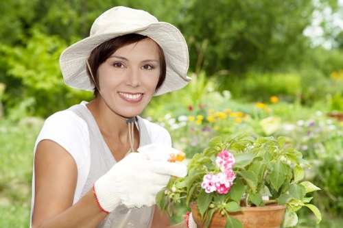 The image depicts a woman in a well-maintained garden during daytime, surrounded by lush greenery, flowering plants, and a neatly trimmed lawn. She is wearing a wide-brimmed hat, white gardening gloves, and a light-colored shirt, and is gently tending to a potted plant with pink and white flowers. The garden features a mix of natural grass, flowering bushes, and small trees, with a paved pathway or patio area in the background, indicative of an outdoor landscaped space suitable for gardening and outdoor leisure. The lighting suggests a mild, possibly overcast day with soft, diffuse light highlighting the vibrant greens and floral colours, creating a calm and inviting environment. Such a garden setup reflects professional landscaping and gardening practices, akin to those supported by companies like Landscapers Ilford, serving residential properties in the Ilford area and surrounding postcode regions. The scene captures an outdoor space that combines aesthetic appeal with functional gardening features, perfect for enhancing home gardens and outdoor living areas.
