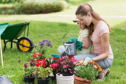 In a well-maintained garden in Ilford, two women, likely a mother and daughter, are engaged in planting and tending to colourful flowering plants in a small outdoor space. The garden features a lush, green lawn with dense grass, bordered by flower beds filled with vibrant red, pink, purple, and orange blooms. In the background, there are trees and a neatly trimmed hedge creating a natural boundary, while a wheelbarrow with black and yellow components is positioned on the grass, suggesting ongoing gardening activity. The women are using watering cans and garden tools, focused on nurturing the plants, with one wearing gardening gloves. The lighting is natural, indicating a bright, possibly sunny day. The scene captures a peaceful and productive moment in a landscaped garden, exemplifying outdoor maintenance and gardening services that a professional landscaper in Ilford might provide, including plant care and garden enhancement to ensure healthy growth and aesthetic appeal.