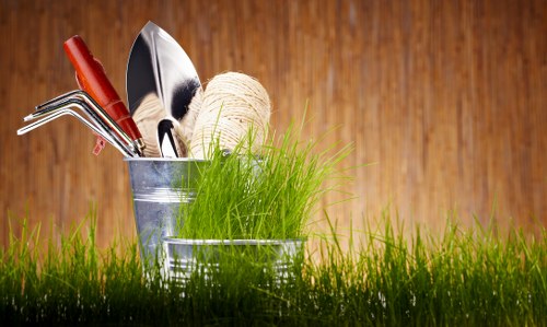 A metal gardening bucket filled with various hand tools, including a small trowel, a cultivator, and a fork, is placed on a lush, green lawn with dense, healthy grass blades. Next to the bucket, there is a ball of natural twine and a bundle of fresh, bright green grass or seedlings. The background features a warm wooden fence or shed, providing a natural backdrop that complements outdoor gardening activities. The scene is set outdoors on a clear day, with even lighting highlighting the textures of the tools, grass, and wood, suggesting preparedness for gardening or landscaping work typical of outdoor spaces in Ilford or surrounding areas. This arrangement reflects professional gardening supplies used by landscapers such as Landscapers Ilford and supports an environment focused on planting, lawn care, and outdoor maintenance, aligning with local gardening services offered in the area. The overall composition emphasizes readiness for planting projects in a typical residential garden setting.