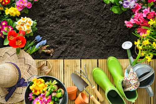 A neatly arranged garden scene viewed from above, featuring a wooden decking area bordered by a lush, green lawn. To the top and sides of the image, vibrant flower beds display a variety of colourful blooms including yellow, pink, purple, and red flowers, adding natural vibrancy. On the decking, garden tools such as a small trowel, a fork, and gardening gloves are neatly placed alongside bright green wellington boots, a watering can, and a decorative floral-patterned sponge, suggesting recent or ongoing planting activities. A wide-brimmed straw hat with a navy ribbon rests near a flower pot filled with pink and yellow blossoms, enhancing the outdoor gardening atmosphere. The background shows rich, dark soil awaiting planting, creating a natural contrast with the vivid greenery and floral displays. Natural daylight illuminates the scene, hinting at a mild, sunny day in Ilford, Essex, suitable for garden maintenance and enhancements. This image effectively captures the essentials of garden planning and planting services provided by Landscapers Ilford, emphasizing healthy lawns, colorful flower beds, and gardening equipment used in creating beautiful outdoor spaces in the local area.