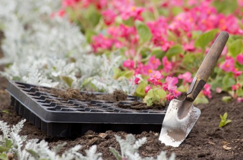 A close-up view of a garden bed in a landscaped outdoor space with dark, rich soil prepared for planting, bordered by a mix of ornamental plants with silver-grey foliage and blooming pink flowers. A small metal gardening trowel with a wooden handle is partially inserted into the soil, indicating recent garden maintenance or planting activity. The background features additional plants and a neatly arranged garden layout, suggesting professional gardening or landscaping work typical of a garden in Ilford, east London. The scene reflects a tidy and thoughtfully designed garden space suitable for ongoing lawn care and outdoor landscaping services by companies such as Landscapers Ilford, with natural light illuminating the vibrant colours and textures of the plants and soil.