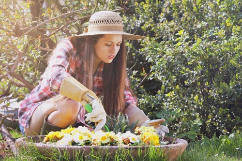 A young woman wearing a wide-brimmed straw hat, plaid shirt, and gardening gloves is kneeling on a lush green lawn in a garden area, carefully tending to a bed of yellow, white, and purple flowers. The garden features a well-maintained grassy lawn, bordered by a variety of dense, leafy shrubs and small trees in the background, providing a natural, vibrant setting typical of outdoor landscape spaces in Ilford. The sunlight filters through the foliage, suggesting a bright, clear day, with the scene capturing a moment of peaceful gardening activity. This outdoor environment showcases typical elements of a landscaped residential garden, including flower beds with defined borders, a mixture of plant textures and colors, and a naturalistic layout that could be part of professional gardening or landscaping services offered by companies in the area. The woman is focused on her planting task, contributing to the overall aesthetics and health of the garden, aligning with gardening services that enhance outdoor beauty and maintain healthy landscape features in Ilford or surrounding areas.