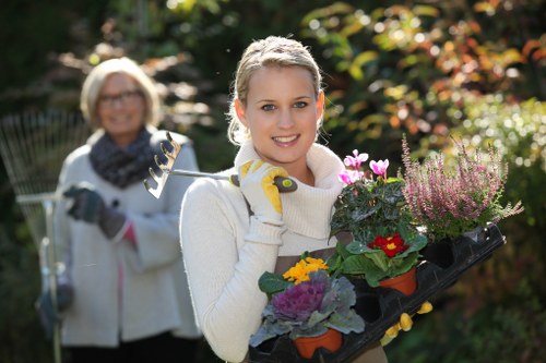A young woman with blonde hair tied back, smiling at the camera while holding a black tray filled with various flowering plants and shrubs in a well-maintained garden. She is wearing gardening gloves and a light-colored sweater, standing outdoors during daylight with a lush, green garden background that includes a variety of plants, trees, and shrubs with a mix of green and reddish foliage. In the background, an older woman with short blonde hair and glasses, dressed in gardening attire, is slightly out of focus, holding a garden rake and working amidst the greenery. The garden features neatly trimmed hedges, flower beds with colorful blooms, and a paved or gravel pathway, suggesting a professionally landscaped outdoor space typical of residential gardens in Ilford or nearby areas, with sunlight filtering through the trees creating a natural, vibrant atmosphere.